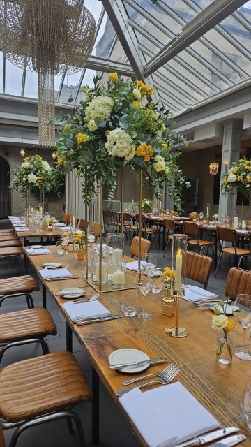 Banquette tablescape design at hampton manor for Mediterranean Shipping Company - Tall gold stands with large yellow and ivory flowers displays - short stone urns of flowers with gold and glass bud vases of flowers and gold and glass candle holders in between