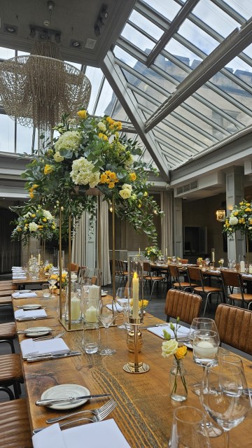 Banquette tablescape design at hampton manor for Mediterranean Shipping Company - Tall gold stands with large yellow and ivory flowers displays - short stone urns of flowers with gold and glass bud vases of flowers and gold and glass candle holders in between