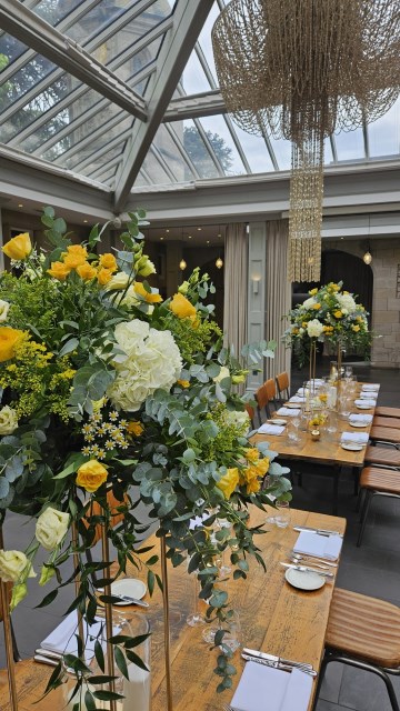 Banquette tablescape design at hampton manor for Mediterranean Shipping Company - Tall gold stands with large yellow and ivory flowers displays - short stone urns of flowers with gold and glass bud vases of flowers and gold and glass candle holders in between