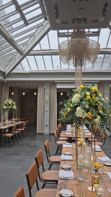 Banquette tablescape design at hampton manor for Mediterranean Shipping Company - Tall gold stands with large yellow and ivory flowers displays - short stone urns of flowers with gold and glass bud vases of flowers and gold and glass candle holders in between