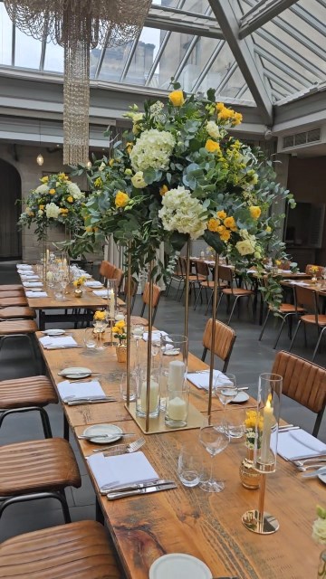 Banquette tablescape design at hampton manor for Mediterranean Shipping Company - Tall gold stands with large yellow and ivory flowers displays - short stone urns of flowers with gold and glass bud vases of flowers and gold and glass candle holders in between