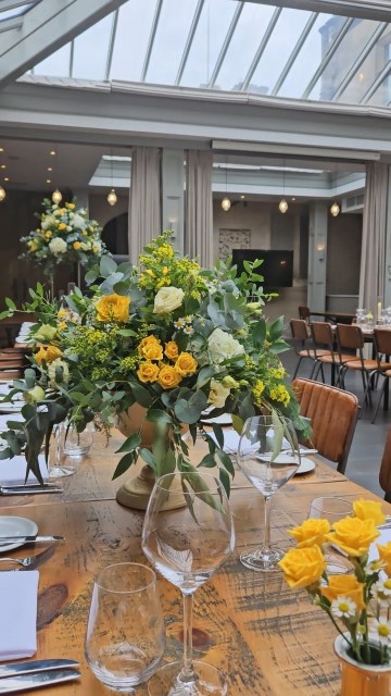 Banquette tablescape design at hampton manor for Mediterranean Shipping Company - Tall gold stands with large yellow and ivory flowers displays - short stone urns of flowers with gold and glass bud vases of flowers and gold and glass candle holders in between